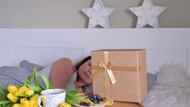 Una giovane donna si svegliò a letto e vide la colazione a letto, fiori e una scatola con un regalo. Sorpresa per mamma e moglie nel giorno delle donne. — Video Stock