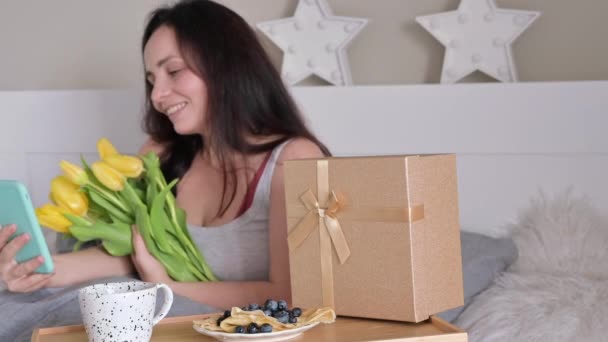Yatakta genç bir kadın akıllı telefondan video ile konuşuyor. Yatakta romantik bir araba, çiçekler ve hediyeli bir kutu. Kadınlar Günü 'nde sevgili karın ve annen için bir sürpriz.. — Stok video