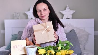 8 Mart, Kadınlar Günü 'nde anneme hediye. Anneler Günü. Elinde sürpriz bir kutu, çiçekler ve yatakta kahvaltı tutan güzel esmer kadın..