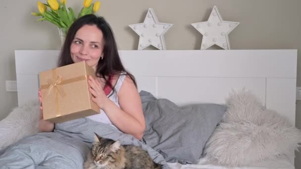 Ung vacker kvinna hålla en låda med en båge och en katt sitter bredvid den. En gåva och en bukett blommor till din älskade mor och hustru på morgonen i sängen. Överraskning den 8 mars — Stockvideo