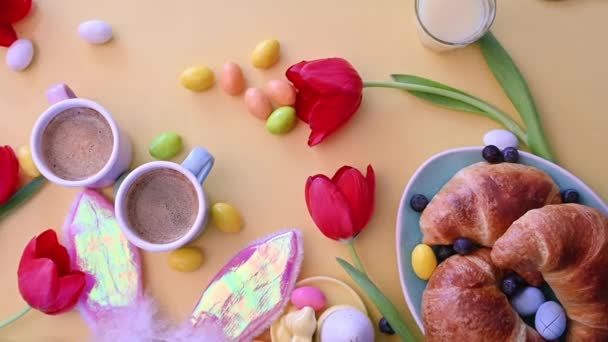 Café expresso em xícaras brilhantes, ovos de chocolate de Páscoa, croissants, orelhas de coelho rosa e tulipas vermelhas em um fundo amarelo. — Vídeo de Stock