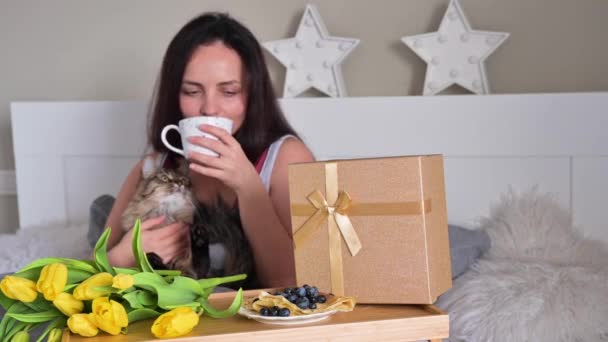 Młoda kobieta otrzymała rano niespodziankę w łóżku, jest szczęśliwa i przytula kota. Kawa, śniadanie i bukiet kwiatów dla dziewczyny w Dzień Kobiet, 8 marca. Niespodzianka dla twojej ukochanej.. — Wideo stockowe