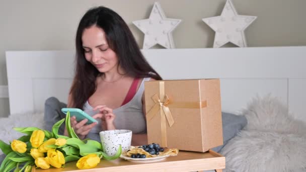 En ung kvinna fick en överraskning på morgonen i sängen, hon ringer i telefon och är glad. Kaffe, frukost och en bukett blommor för en flicka på Womens Day, 8 mars. — Stockvideo