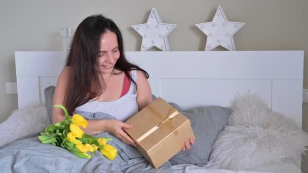 En gåva och en bukett blommor till din älskade mor och hustru på morgonen i sängen. Ung vacker kvinna hålla en låda med en båge och en katt sitter bredvid den. Överraskning den 8 mars. — Stockvideo