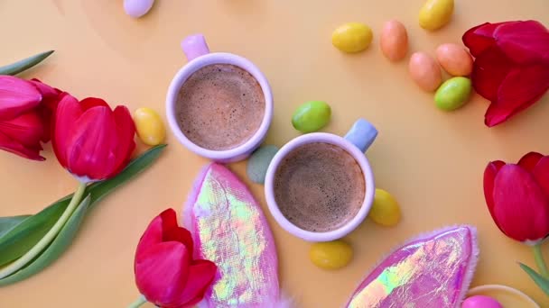 Twee kopjes geurige espresso en chocolade paaseieren op een gele achtergrond. Bloemen, gemaskerde konijnenoren, zwarte koffie en snoep. Zicht van bovenaf. Paasvakantie — Stockvideo