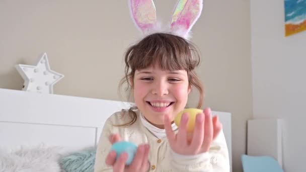 Malá šťastná holka v králičích uších se baví, hraje si s barevnými velikonočními vejci. Čokoládová vejce různých barev na Velikonoce v rukou dítěte. — Stock video