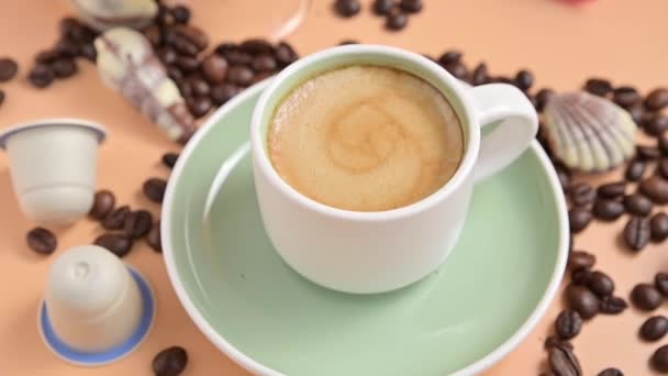 Descanso. Una taza de café expreso aromático con espuma. Bebida tradicional italiana de desayuno sobre un fondo brillante con cápsulas y granos de café. — Vídeos de Stock