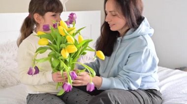 Küçük bir kız annesine bir buket sarı ve mor lale verir. Anneler Günü 'nde anne ve kızının sevgi dolu kucaklaşmaları. Annen için 8 Mart hediyesi..