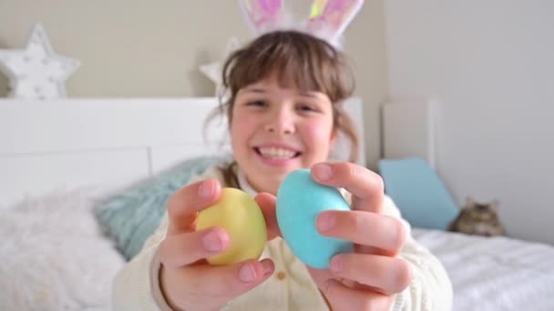 Το μικρό χαρούμενο κορίτσι με αυτιά κουνελιού διασκεδάζει, παίζοντας με χρωματιστά πασχαλινά αυγά. Αυγά σοκολάτας διαφόρων χρωμάτων για το Πάσχα στα χέρια ενός παιδιού. — Αρχείο Βίντεο