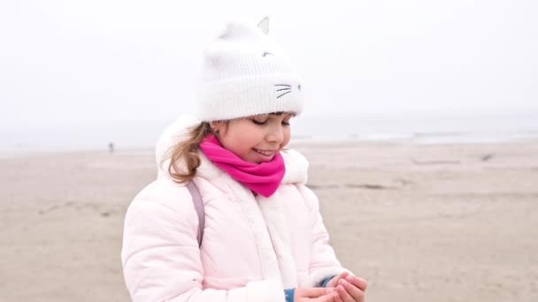 Mała szczęśliwa dziewczynka o europejskim wyglądzie spaceruje wzdłuż nabrzeża morza lub oceanu wczesną wiosną, bawi się muszelkami. Koncepcja rodzinnej rekreacji na świeżym powietrzu. Spowolnienie — Wideo stockowe