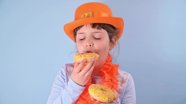 คิงส์เดย์ในฮอลแลนด์ เทศกาลดั้งเดิมในวันที่ 27 เมษายนในเนเธอร์แลนด์ สาวน้อยในหมวกสีส้มฉลองบนพื้นหลังสีฟ้า กินโดนัทสี . — วีดีโอสต็อก