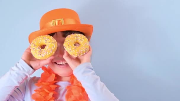 Голландский праздник День короля весной. Традиционный фестиваль 27 апреля в Нидерландах. Маленькая девочка в праздничной оранжевой шляпе на синем фоне ест цветные пончики. — стоковое видео