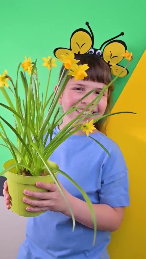 Ein kleines Mädchen versteckte sich hinter gelben Narzissen vor grünem Hintergrund. Glückliches Kind mit Bienenkappen hält einen Topf mit Gartenblumen in der Hand. Pflanzzeit im Garten im Frühling. Selektiver Fokus — Stockvideo