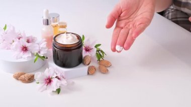 Bir kadın ellerine krem sürer. Badem yağı ve vitaminli el kremi. Vücut sağlığı ve güzellik. Parlak bir arkaplan, kozmetik ve doğal badem çiçekleriyle dolu kavanozlar..