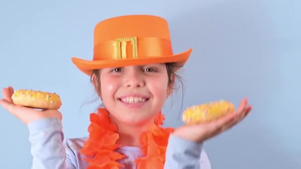 Święto Holenderskie Dzień Króla wiosną. Mała dziewczynka w pomarańczowym kapeluszu na niebieskim tle je kolorowe pączki. Tradycyjny festiwal 27 kwietnia w Holandii. — Wideo stockowe