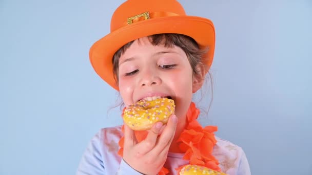 Festival tradicional em 27 de abril na Holanda. Dia do Rei feriado holandês. Uma menina em um boné festivo de carnaval de cor laranja em um fundo azul come donuts coloridos. — Vídeo de Stock