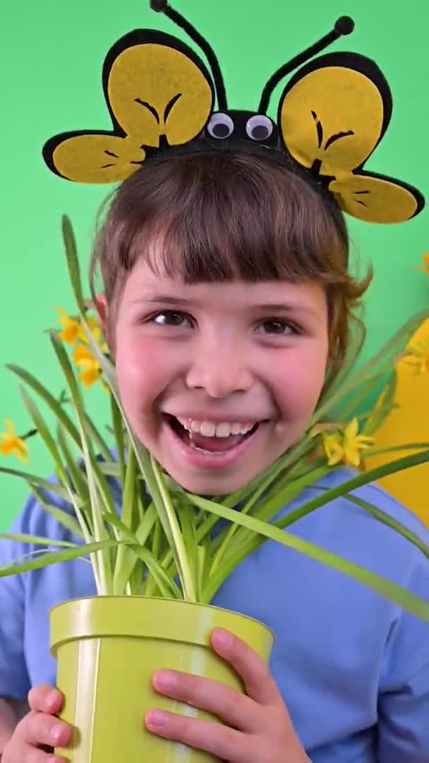 Malá holčička se schovala za žluté narcisy na zeleném pozadí. Šťastné dítě s včelí čepicí drží květináč zahradních květin. Sezóna výsadby v zahradě na jaře. Selektivní zaměření — Stock video