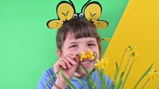 Malá holčička se schovala za žluté narcisy na zeleném pozadí. Šťastné dítě s včelí čepicí drží květináč zahradních květin. Sezóna výsadby v zahradě na jaře. Selektivní zaměření — Stock video