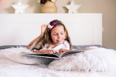Küçük kız parlak bir odada yatağın üstünde kitap okuyor. Çocuk elinde bir kitapla yatak odasında yatıyor ve hevesle kitabı okuyor. Metin için boş alan