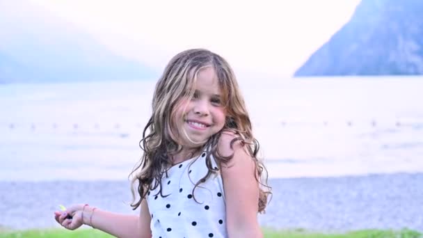 Niña feliz exuda emociones de alegría, ella está girando jugando y sonriendo. Un niño en la orilla de un lago de montaña pasa tiempo con su familia en el verano. Concepto de Felices Fiestas con Padres. — Vídeos de Stock