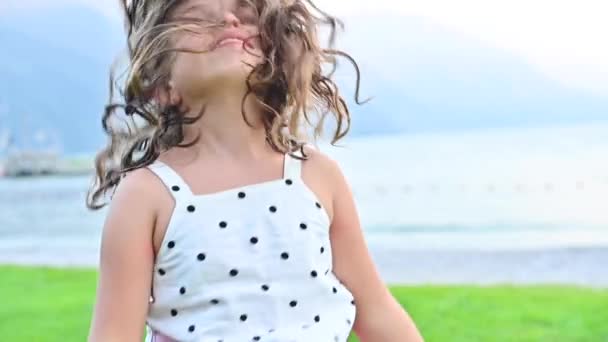 Szczęśliwa dziewczynka emanuje emocjami radości, wiruje grając i uśmiechając się. Dziecko na brzegu górskiego jeziora spędza lato ze swoją rodziną. Wesołych świąt koncepcja z rodzicami. — Wideo stockowe