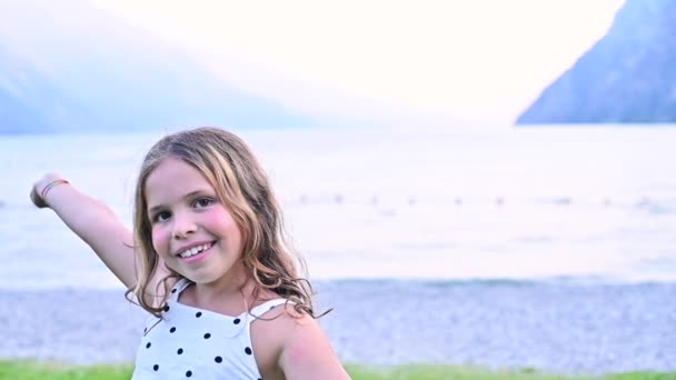 Щаслива маленька дівчинка викриває емоції радості, вона крутиться граючись і посміхаючись. Дитина на березі гірського озера проводить час зі своєю сім'єю влітку. Концепція щасливих свят з батьками . — стокове відео