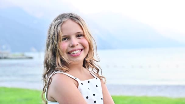 Fröhliches kleines Mädchen strahlt Emotionen der Freude aus, sie dreht sich spielend und lächelt. Ein Kind am Ufer eines Bergsees verbringt im Sommer Zeit mit seiner Familie. Frohe Festtage mit den Eltern. — Stockvideo