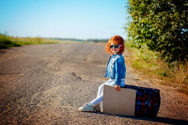 Hübsche kleine lockige Mädchen sitzt auf einem Koffer auf der Straße und wartet auf einen Bus oder ein Auto. Sommerzeit. Reise- und Urlaubskonzept — Stockfoto