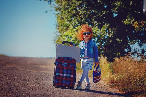 Little curly girl with big suitcase standing on road and waiting a bus or car. — Stock Photo, Image