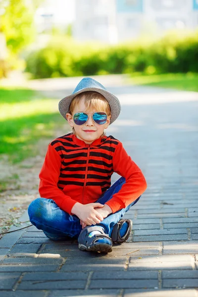 Fashionable little boy in sunglasses and cap sitting on acphalt — Stock Photo, Image