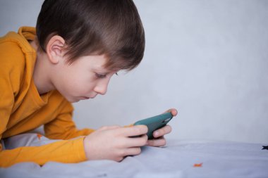 Akıllı telefonlara odaklanmış yatakta uzanan sevimli küçük çocuk. Çocuk akıllı telefonuyla video oyununda oynuyor. Çocuk video oyunu oynarken eğleniyor. Sosyal ve teknoloji kavramı