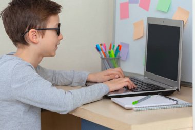 Masada oturan okul çocuğu dizüstü bilgisayarla ödev yapıyor. Çocuklar evde uzaktan öğreniyor. Çocuk internet eğitimi.