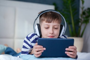 Oturma odasındaki yatakta dijital tabletle oynayan kulaklıklı sevimli çocuk. Eğitim, boş zaman, teknoloji ve internet kavramı
