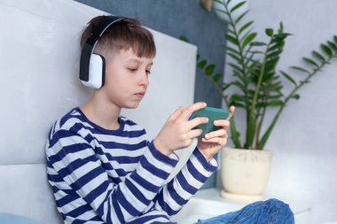 Kulaklıklı çocuk bakıyor ve telefonla oynuyor. Oturma odasında yatakta öğreniyor. Teknoloji kullanan çocuklar.