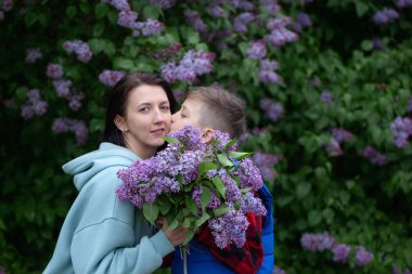 Oğlu annesini öper ve ona bir buket leylak verir. Anne oğul kucaklaşması, anne sevgisi. Anneler Günü.