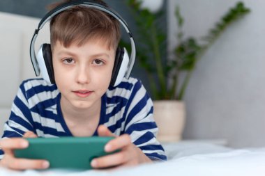 Kulaklıklı şirin çocuk oturma odasındaki yatağın üstünde akıllı telefonuyla oynuyor. Eğitim, boş zaman, teknoloji ve internet kavramı