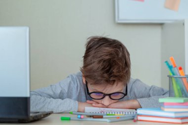 Mutsuz ve gergin bir okul çocuğu evdeki masasında not defteriyle ders çalışıyor. Çocuk çevrimiçi öğrenmekten bıktı. Çocuk yüzüstü masada dinleniyor. Eğitim ve çocukluk kavramı