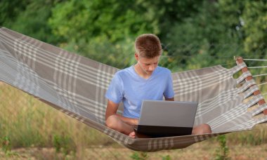 Yaz bahçesindeki hamakta dizüstü bilgisayarı olan sevimli genç bir adam. Uzaktan çalışma, eğitim, serbest meslek, dinlenme ve rahatlama kavramını benimse