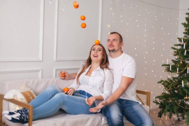 Beyaz gömlekli ve kotlu genç bir çift evde süslü Noel ağacına karşı kanepede mandalinalarla eğleniyor. Konforlu ev konseptinde kış tatili