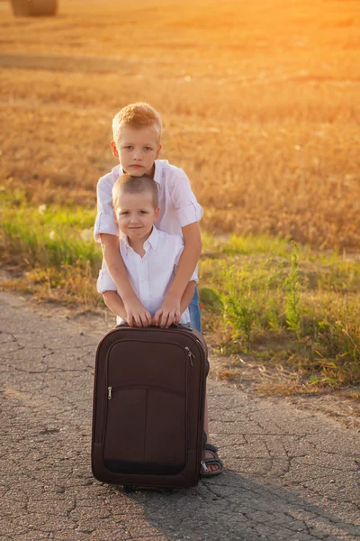 Dos hermanos con una maleta en la carretera en el verano al atardecer — Foto de Stock