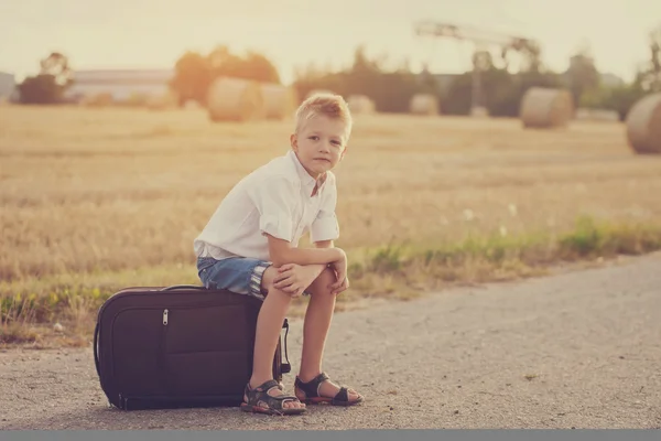 The happy child sits on a suitcase in the summer sunny day, the — Stock Photo, Image