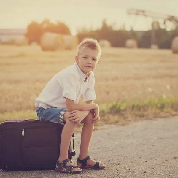 The happy child sits on a suitcase in the summer sunny day, the — Stock Photo, Image