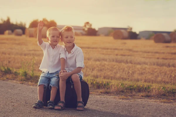 Due fratelli si siedono su una valigia sulla strada in estate al tramonto — Foto Stock