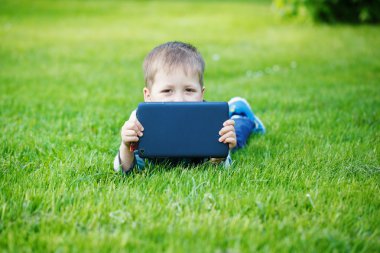 Parkta çimenlerde yatan ve tablet kullanan sevimli çocuk.