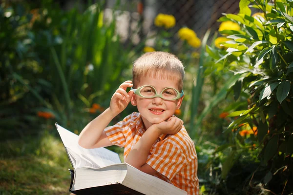 Cute kid boy with glasses, reading a book in garden, sitting on grass. — Stock Photo, Image