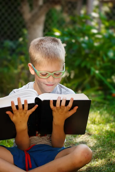 Sød dreng med briller, læser en bog i haven, sidder på græs . - Stock-foto