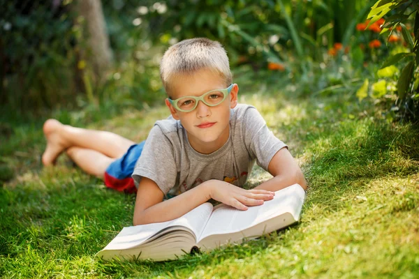 Söt pojke med glasögon ligger på grönt gräs, läsa bok i sommardag. — Stockfoto