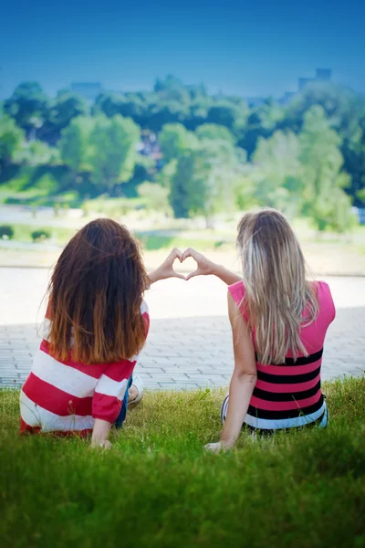 Dwie dziewczyny siedzą na trawie i Pokaż ręce serce, lusterka, pojęcie przyjaźni. Nieostrość na ręce — Zdjęcie stockowe
