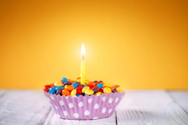 Decorated Birthday cupcake with one lit candle and colorful candies on yellow background. Holidays greeting card