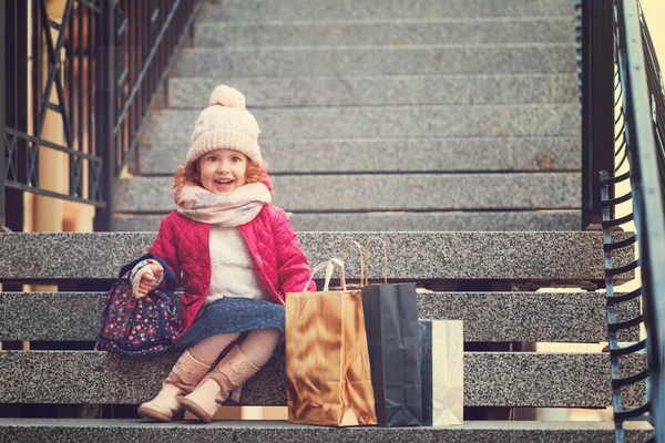Lille pige i hat, jakke og tørklæde holder farverig shoppin - Stock-foto